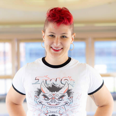 A shot from the bust up of a femme-presenting individual with a bright red wavy mohawk. They are wearing hoop earrings, red lipstick, and a white t-shirt with a fictional oni and the word TWO on it.